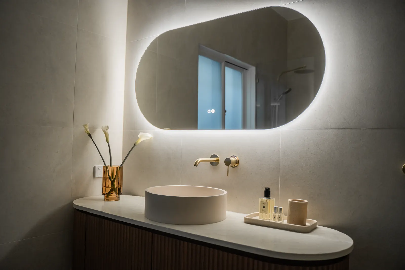 Evening view — backlit mirror glow over walnut vanity and porcelain tiled walls
